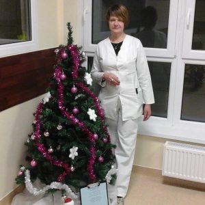 Kambaryje su dideliais langais ir radiatoriumi šalia papuoštos Kalėdų eglutės, papuoštos rožiniais ir baltais papuošalais, stovi žmogus, vilkintis baltą uniformą.