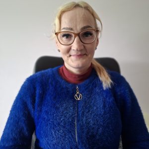 Moteris su šviesiais plaukais, akiniais ir mėlynu pūkuotu megztiniu sėdi ant kėdės, atsukusi veidą į kamerą. Ji šiek tiek šypsosi, o ant kaklo nešioja vėrinį su apvaliu pakabuku.