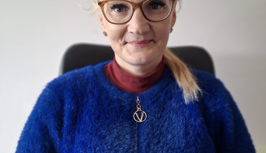 Moteris su šviesiais plaukais, akiniais ir mėlynu pūkuotu megztiniu sėdi ant kėdės, atsukusi veidą į kamerą. Ji šiek tiek šypsosi, o ant kaklo nešioja vėrinį su apvaliu pakabuku.